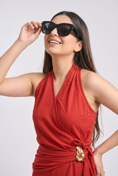 A woman wearing black cat-eye sunglasses and a red halter dress with a gold buckle, posing against a light gray background.