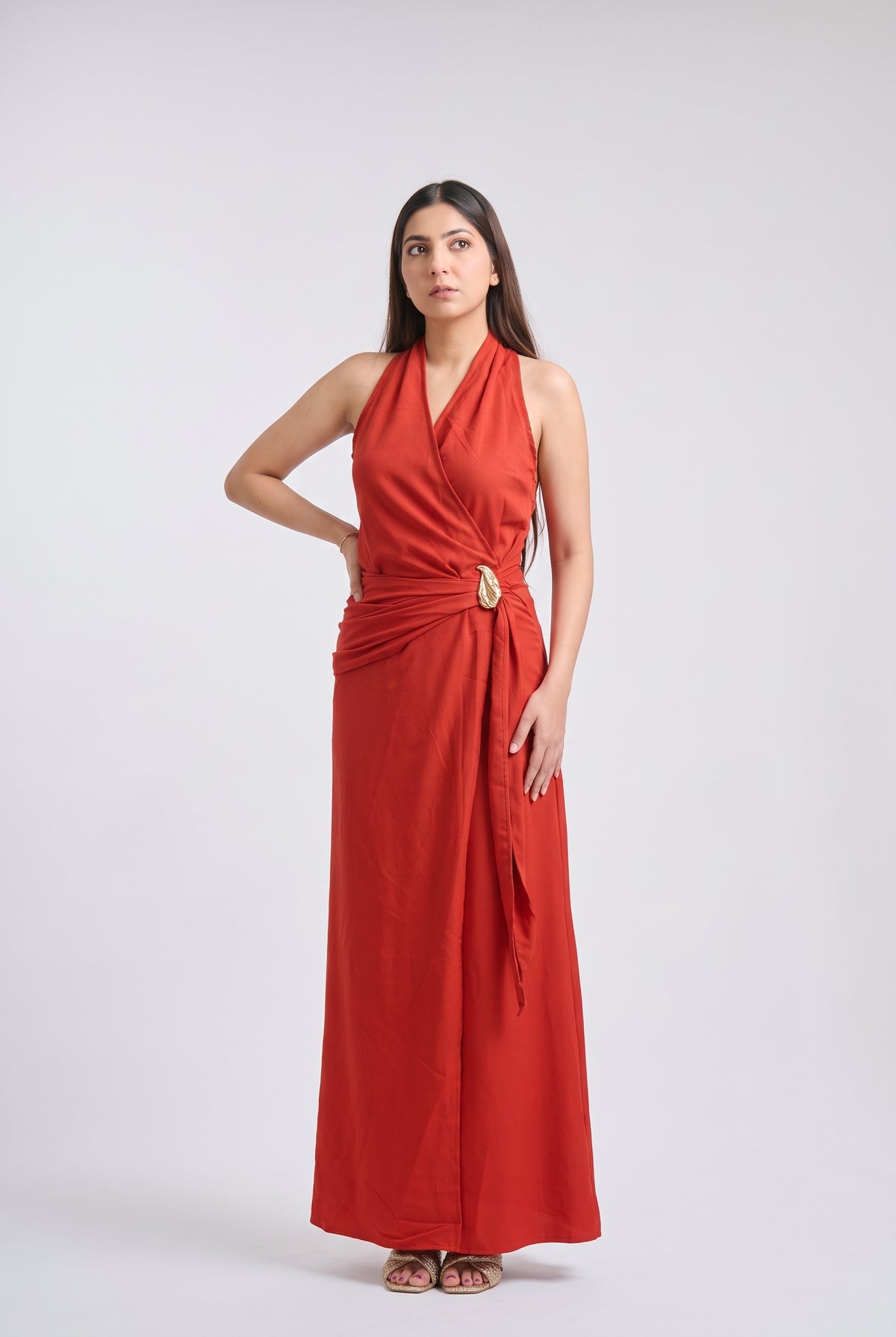 A woman modeling a sleeveless, wrap-style maxi dress in a vibrant rust red color, featuring a draped waist with a gold-tone decorative ring, set against a plain light gray studio background.