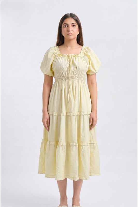 A woman modeling a light yellow tiered midi dress with puff sleeves and a smocked bodice, photographed against a plain light gray studio background.