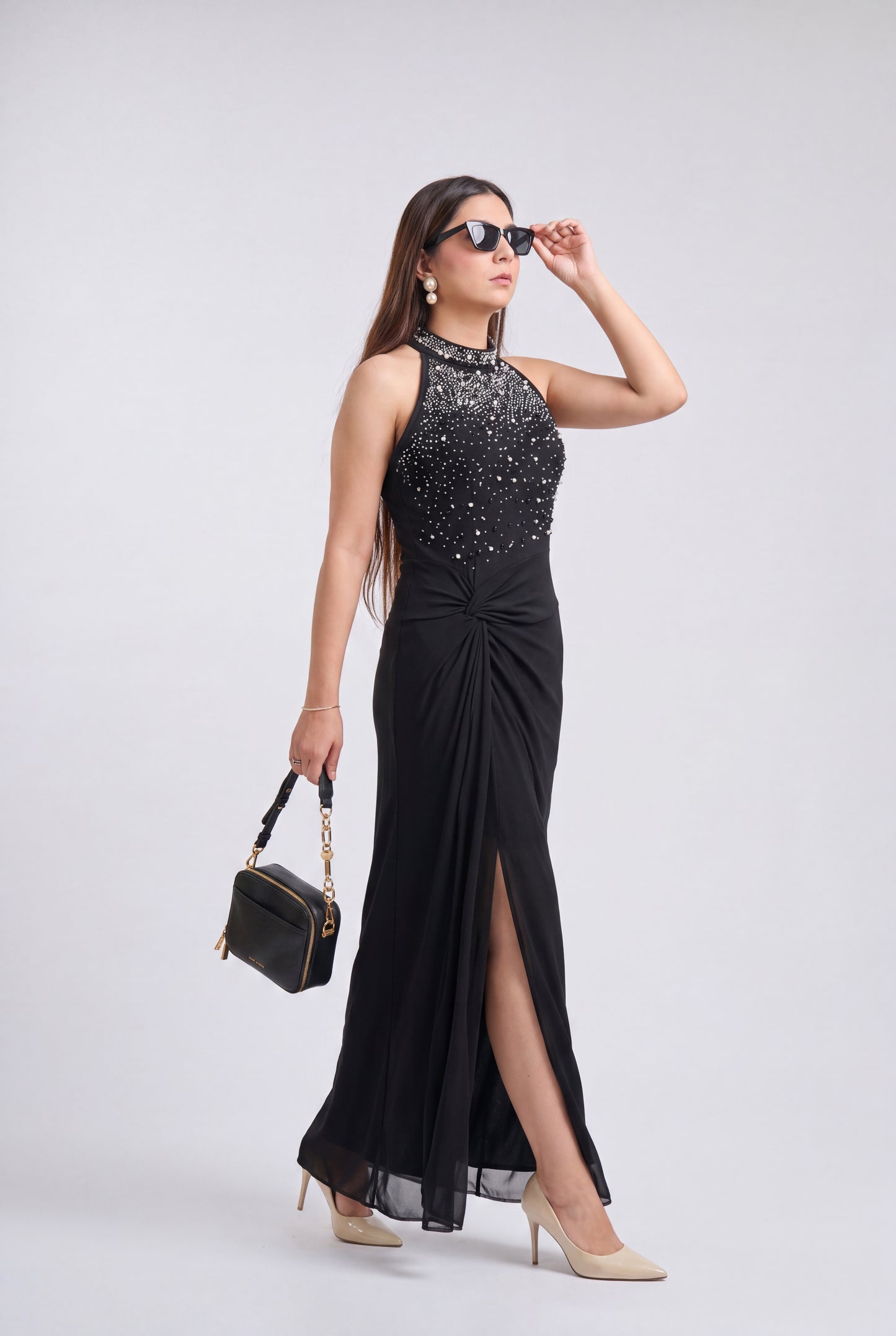 A woman models a sleeveless black evening gown with a high-neck bodice covered in sparkling silver embellishments, a gathered waist, and a high front slit. She is wearing beige heels, holding a small black handbag, and posing against a light gray studio background.