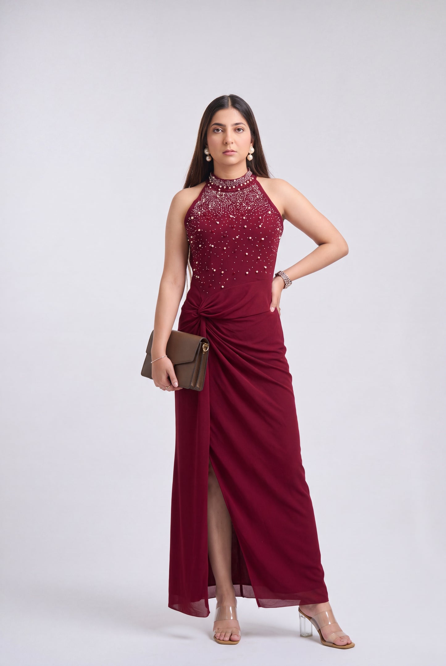 A woman modeling a sleeveless halter-neck maxi dress in deep burgundy with a beaded embellished bodice and a draped skirt, holding a brown clutch against a light gray studio background.