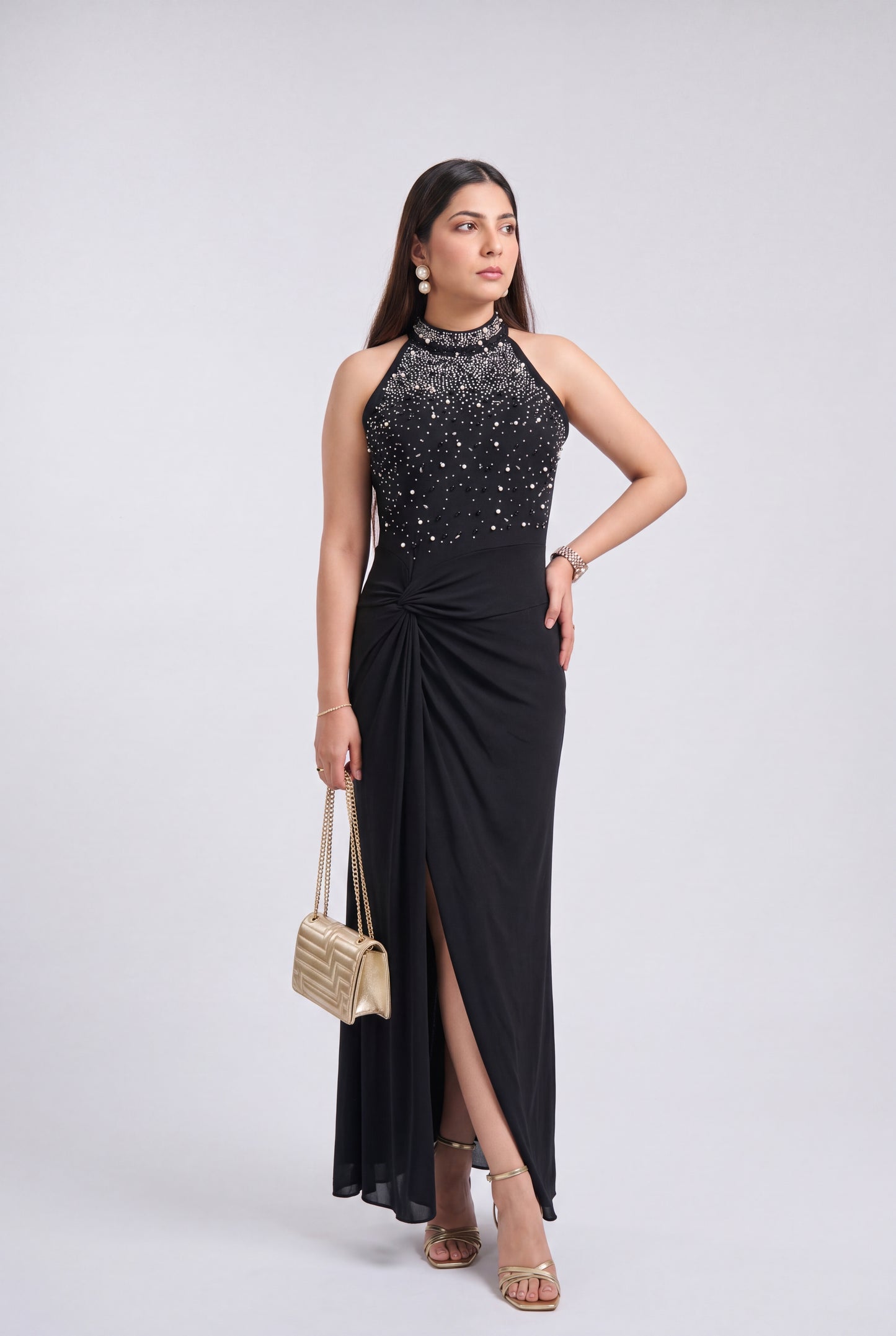 A woman modeling a sleeveless black evening gown with a high-neck beaded bodice, a gathered skirt, and a thigh-high slit, styled with a small woven handbag and metallic sandals against a light gray studio background.