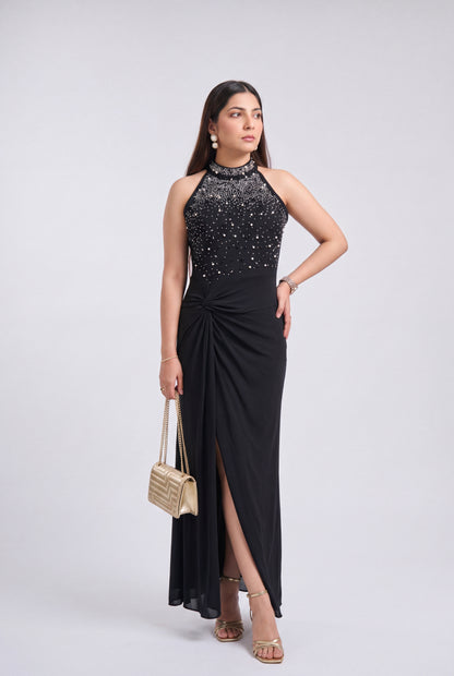 A woman modeling a sleeveless black evening gown with a high-neck beaded bodice, a gathered skirt, and a thigh-high slit, styled with a small woven handbag and metallic sandals against a light gray studio background.