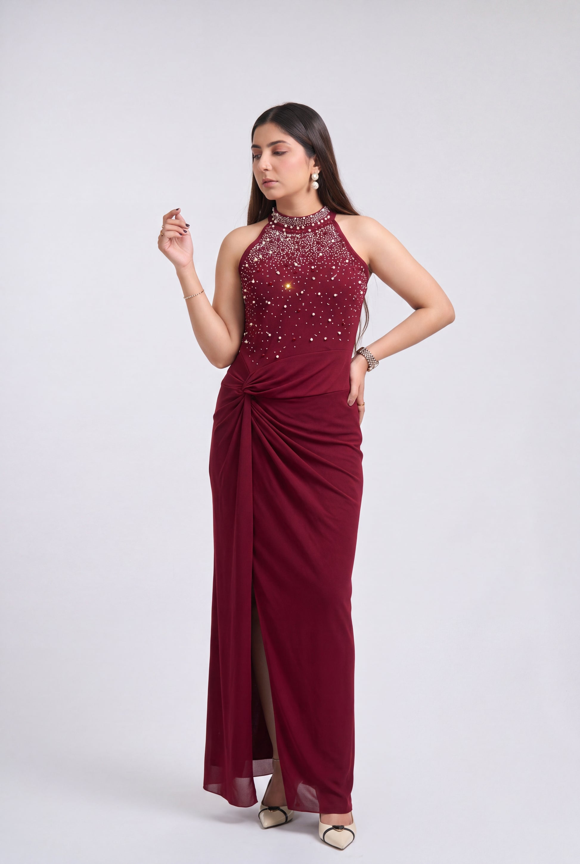 A woman modeling a sleeveless, floor-length maroon evening gown with a high-neck beaded bodice, draped side detail, and a thigh-high slit, photographed against a light gray studio background.