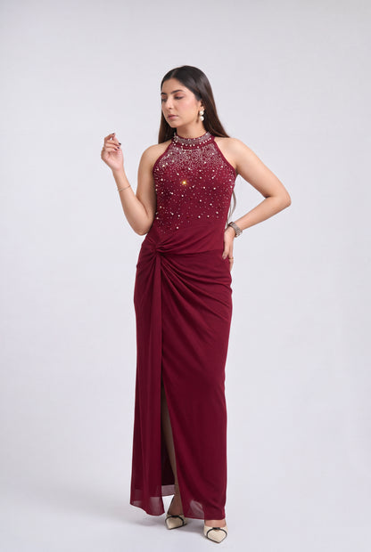 A woman modeling a sleeveless, floor-length maroon evening gown with a high-neck beaded bodice, draped side detail, and a thigh-high slit, photographed against a light gray studio background.