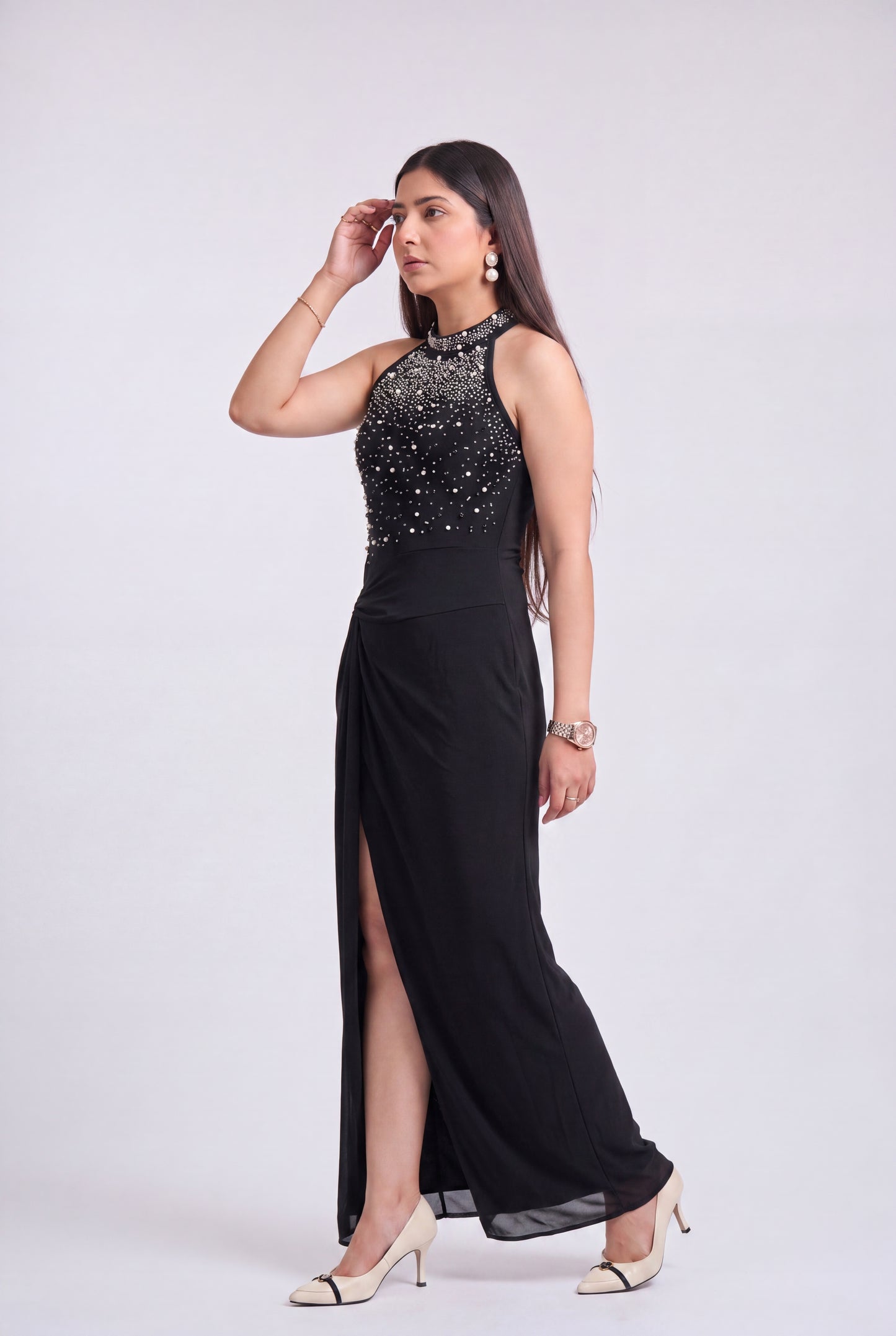 A woman modeling a sleeveless black evening gown with a high-neckline, heavily embellished beaded bodice, and a high side slit, paired with beige heels against a light gray studio background.
