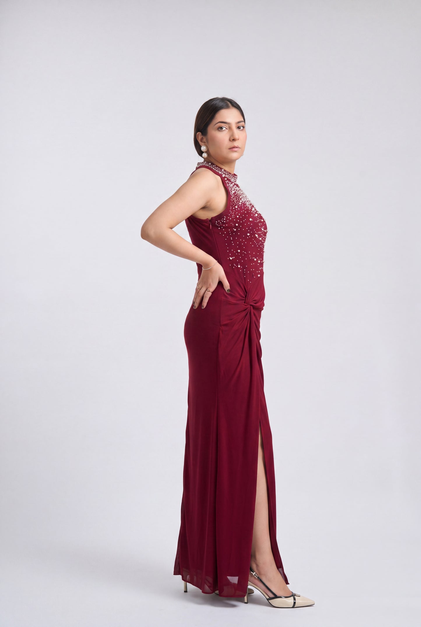 A woman modeling a sleeveless, floor-length burgundy gown with a high neckline, beaded embellishments on the bodice, and a draped side slit, photographed against a light gray studio background.