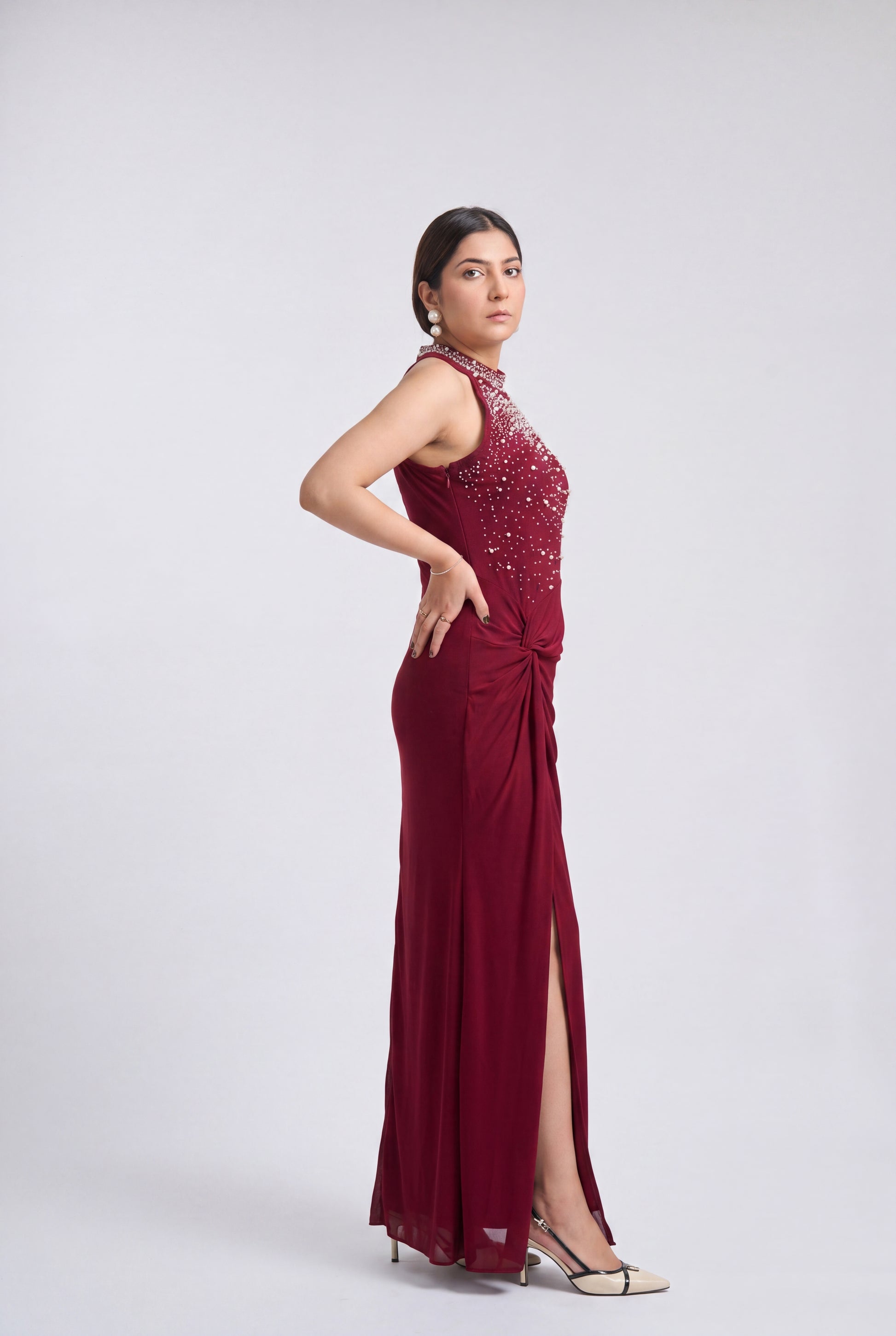 A woman modeling a sleeveless, floor-length burgundy gown with a high neckline, beaded embellishments on the bodice, and a draped side slit, photographed against a light gray studio background.