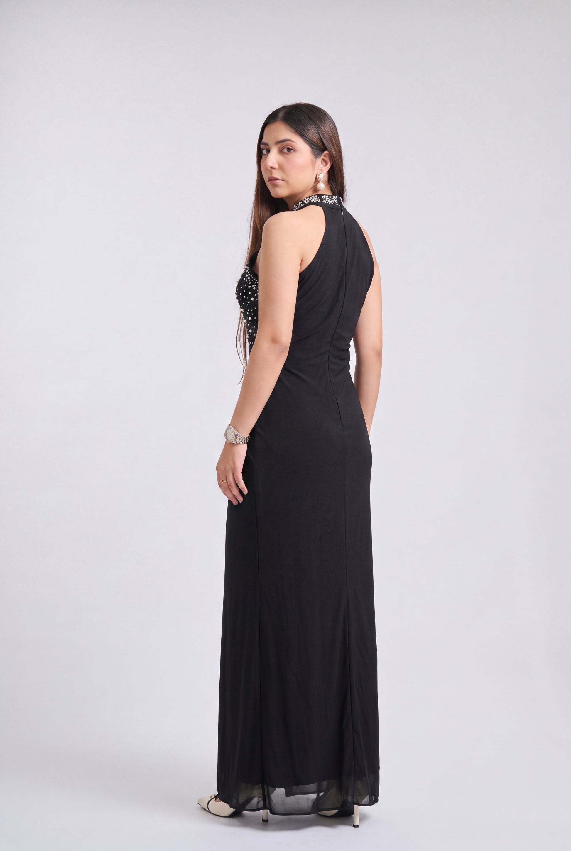 A woman modeling a sleeveless black maxi dress with a beaded embellished bodice, shown from a side-back angle against a light gray studio background.