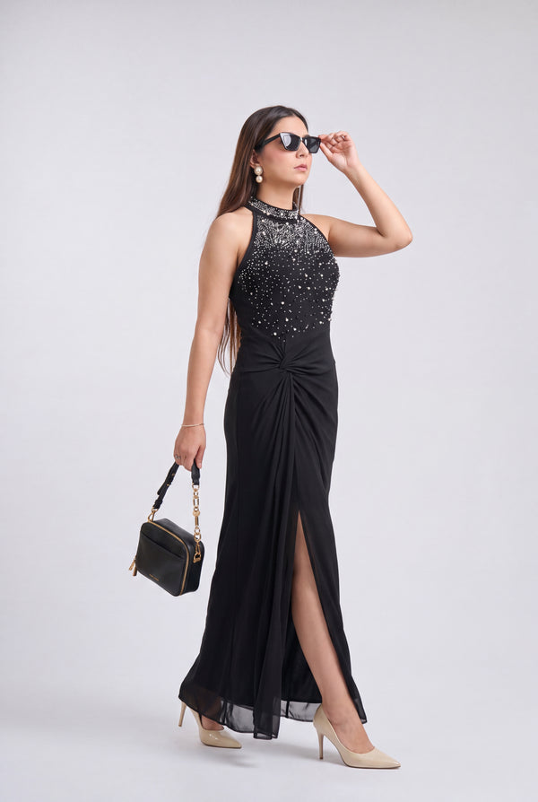 A woman models a sleeveless black evening gown with a high-neck bodice covered in sparkling silver embellishments, a gathered waist, and a high front slit. She is wearing beige heels, holding a small black handbag, and posing against a light gray studio background.