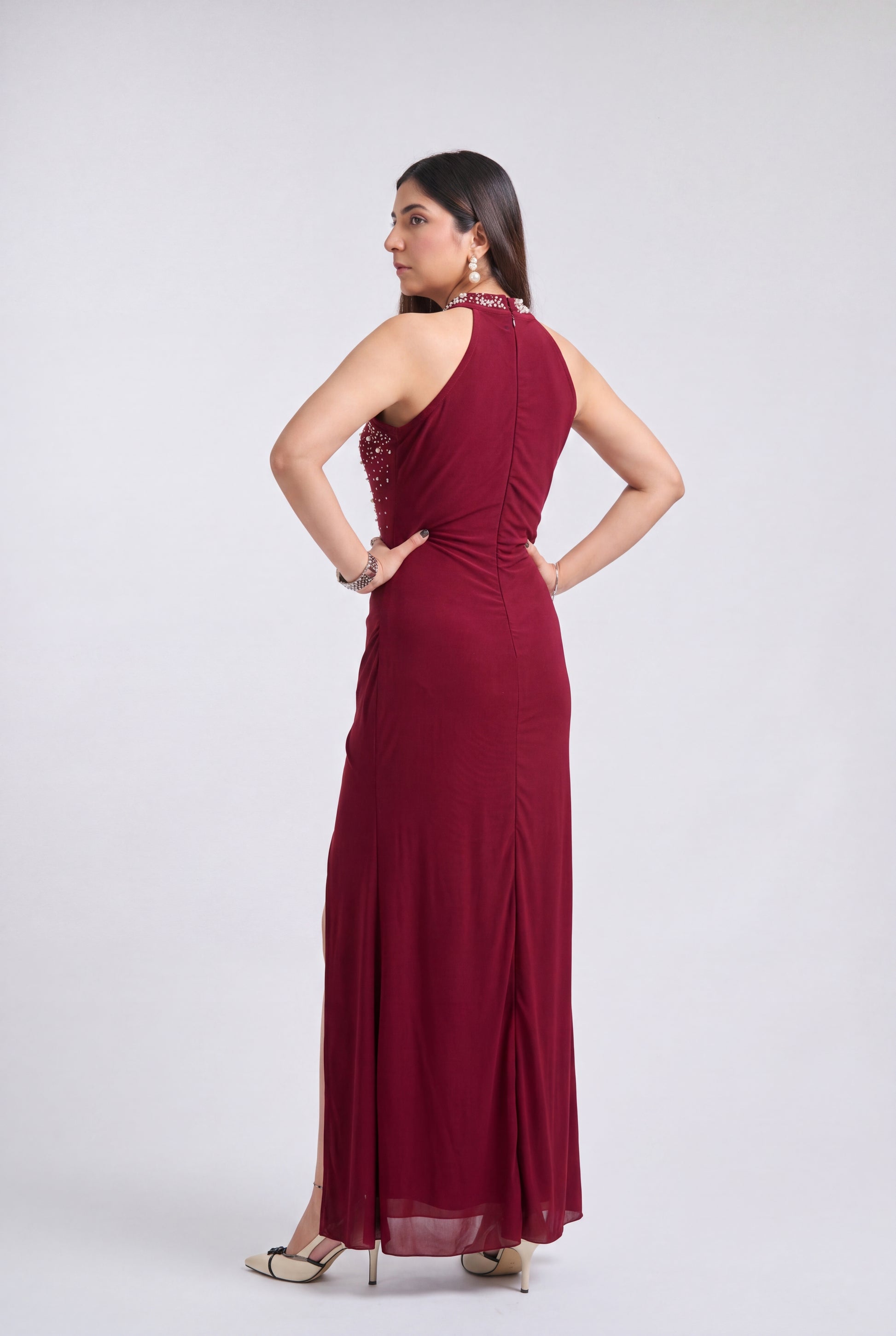 A woman modeling a sleeveless, floor-length burgundy maxi dress from the back, standing against a light gray studio background.