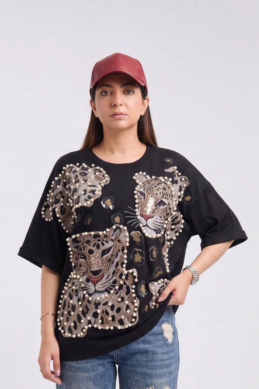 A woman wearing a black oversized t-shirt with large leopard and cheetah prints embellished with gold and white rhinestones, paired with a maroon baseball cap and distressed blue jeans, against a light gray studio background.