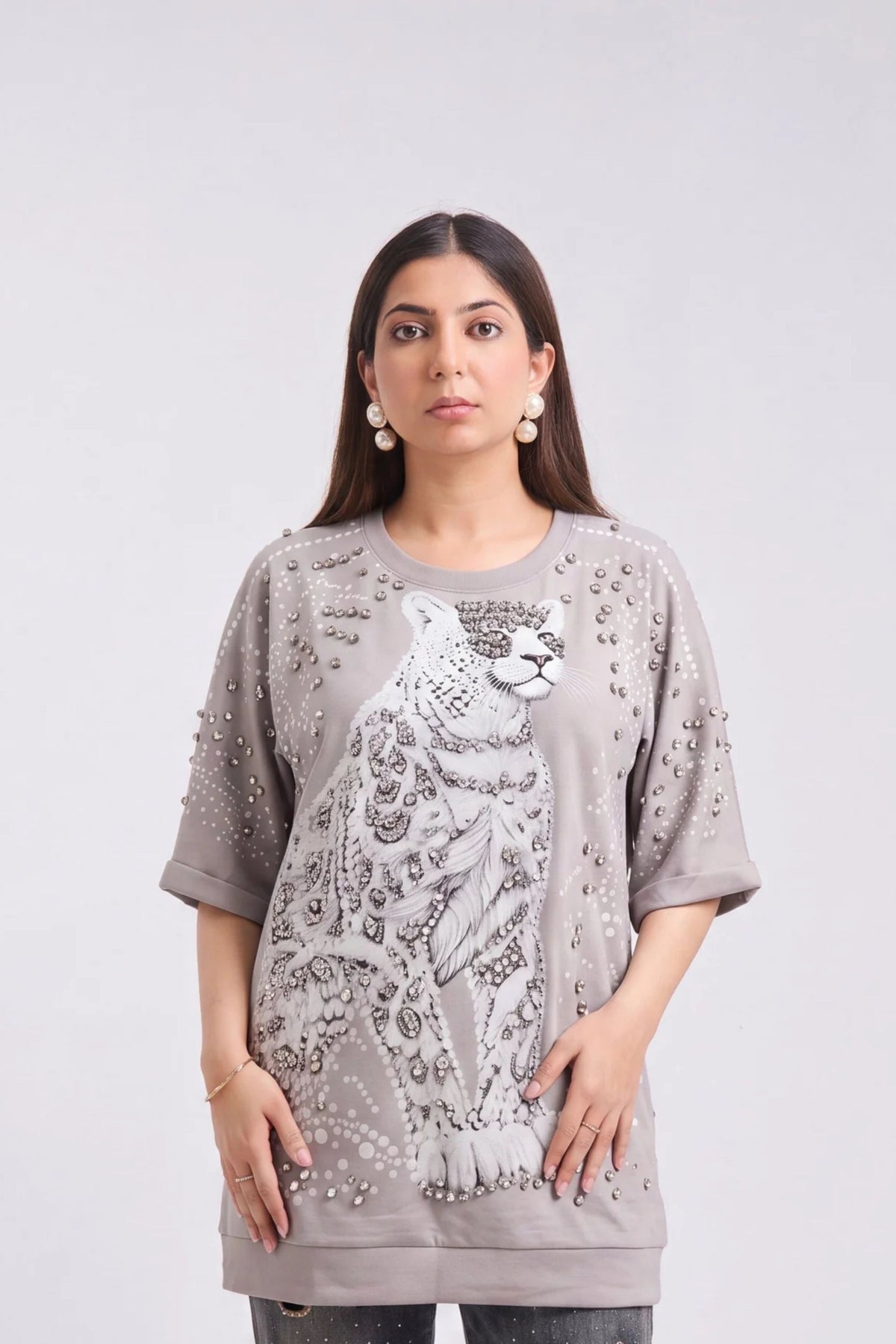 A woman modeling an oversized taupe-gray t-shirt featuring a large white leopard graphic and silver rhinestone embellishments on the front, set against a plain light gray background.