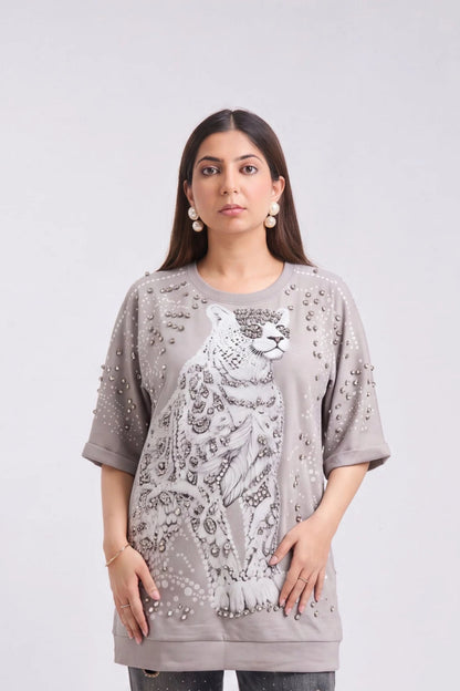A woman modeling an oversized taupe-gray t-shirt featuring a large white leopard graphic and silver rhinestone embellishments on the front, set against a plain light gray background.