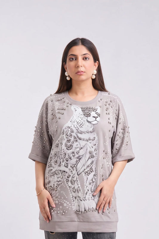 A woman modeling an oversized taupe-gray t-shirt featuring a large white leopard graphic and silver rhinestone embellishments on the front, set against a plain light gray background.
