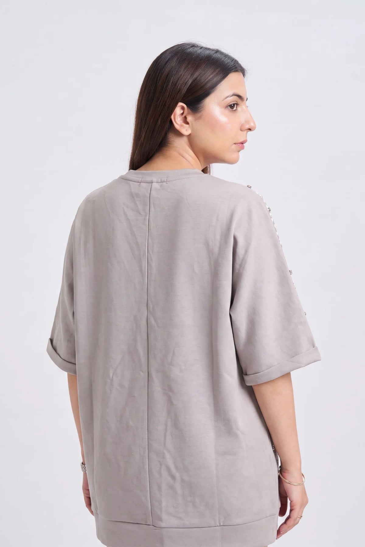 A woman shown from the back wearing a loose, short-sleeve taupe-gray top with rolled cuffs and decorative side embellishments, photographed against a plain light gray studio background.