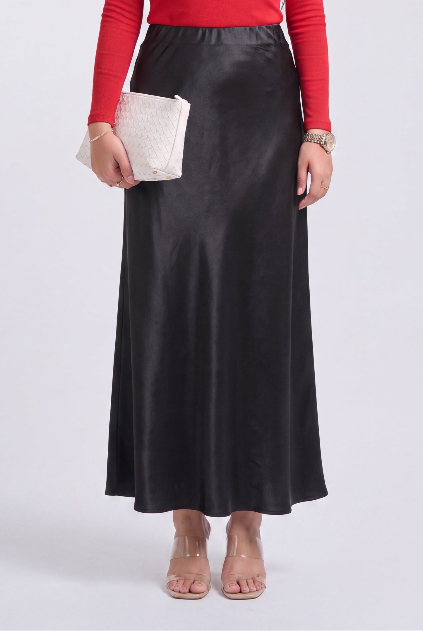 A model wearing a black satin midi skirt, a red long-sleeve top, and clear block-heel sandals, holding a white quilted clutch.