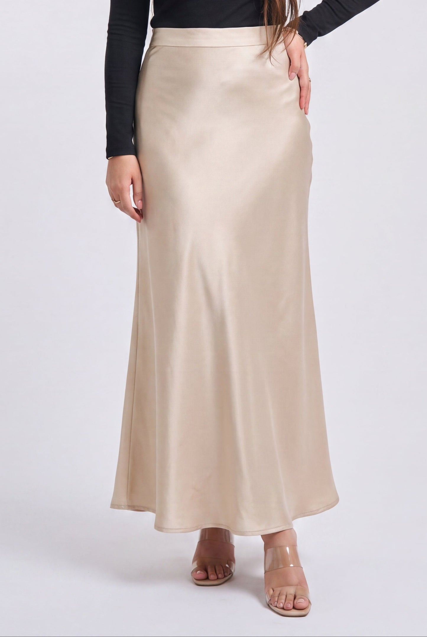 A woman modeling a black long-sleeve top and a high-waisted, floor-length champagne beige satin skirt against a light gray studio background.