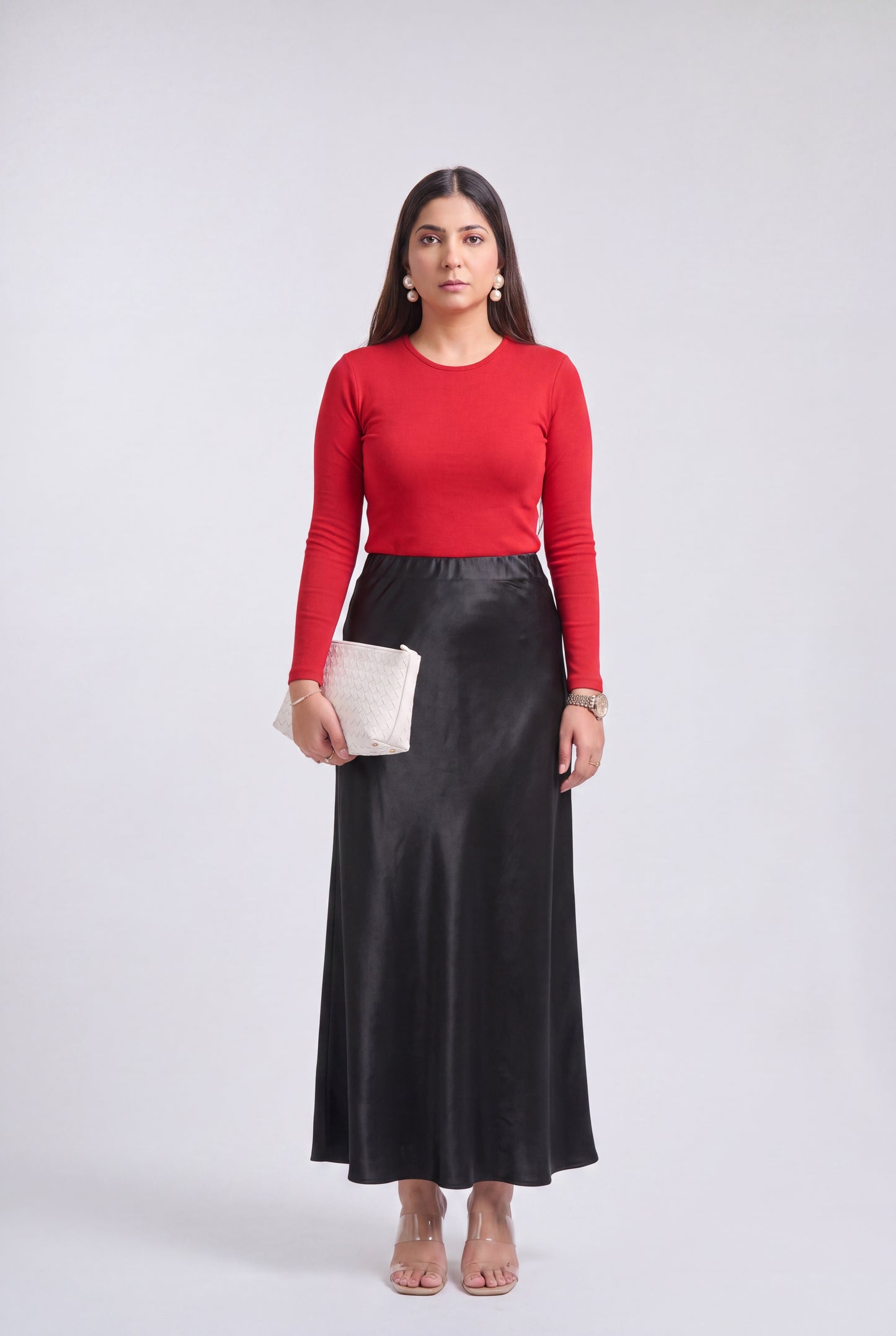 A woman stands against a light gray studio background wearing a bright red long-sleeve knit top and a long black satin-like skirt, holding a small white clutch.