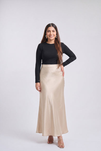 A woman modeling a black long-sleeve top and a high-waisted, floor-length champagne beige satin skirt against a light gray studio background.