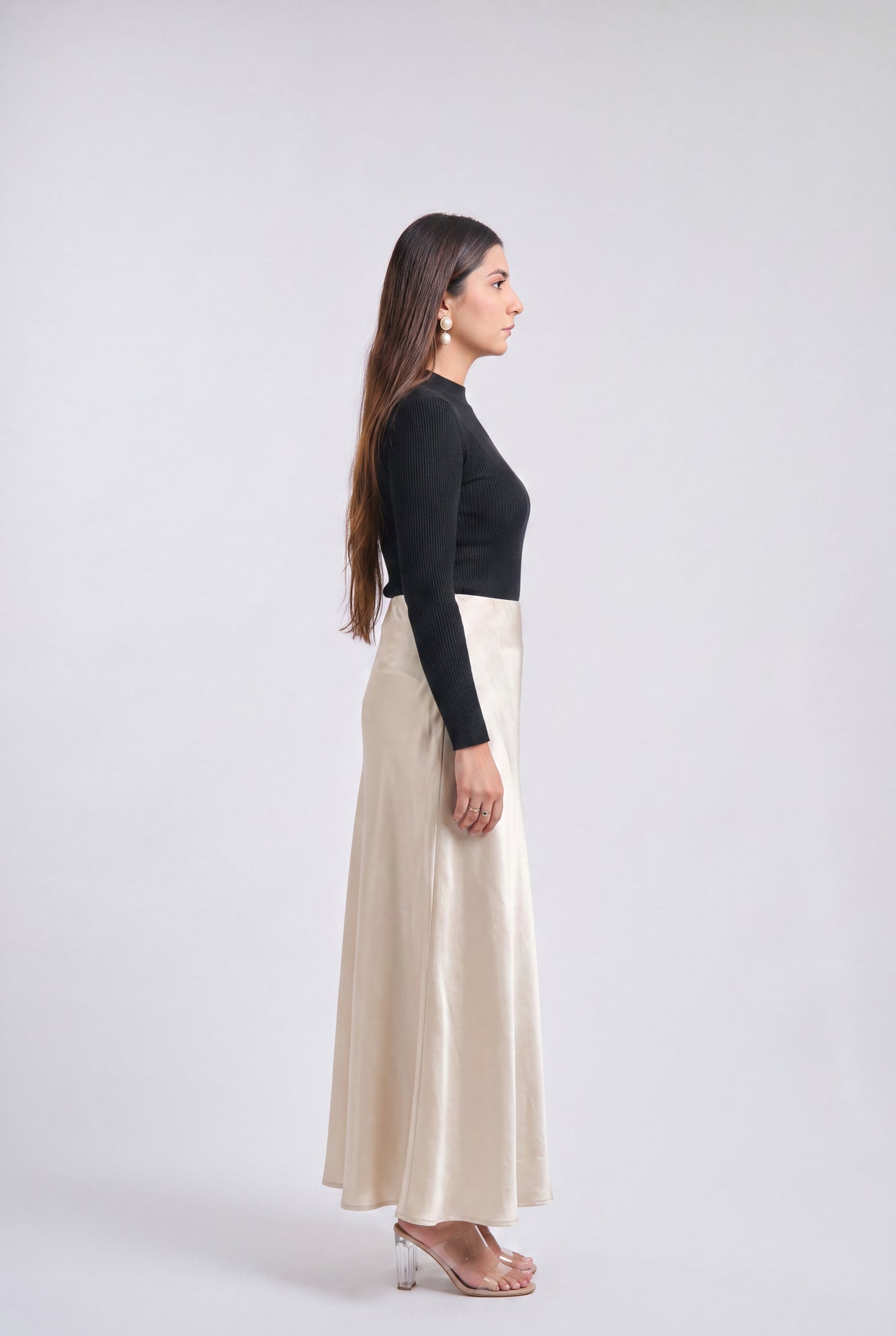 A woman in side profile wearing a black long-sleeve top and a high-waisted, floor-length cream satin skirt, standing against a light gray studio background.