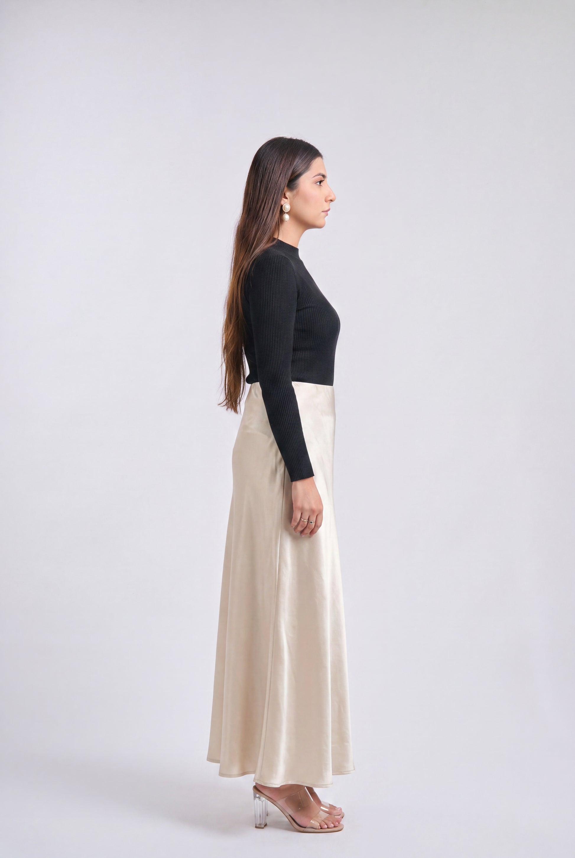 A woman in side profile wearing a black long-sleeve top and a high-waisted, floor-length cream satin skirt, standing against a light gray studio background.