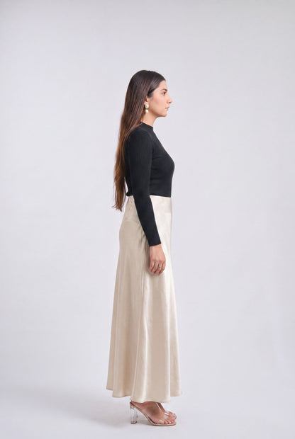 A woman in side profile wearing a black long-sleeve top and a high-waisted, floor-length cream satin skirt, standing against a light gray studio background.
