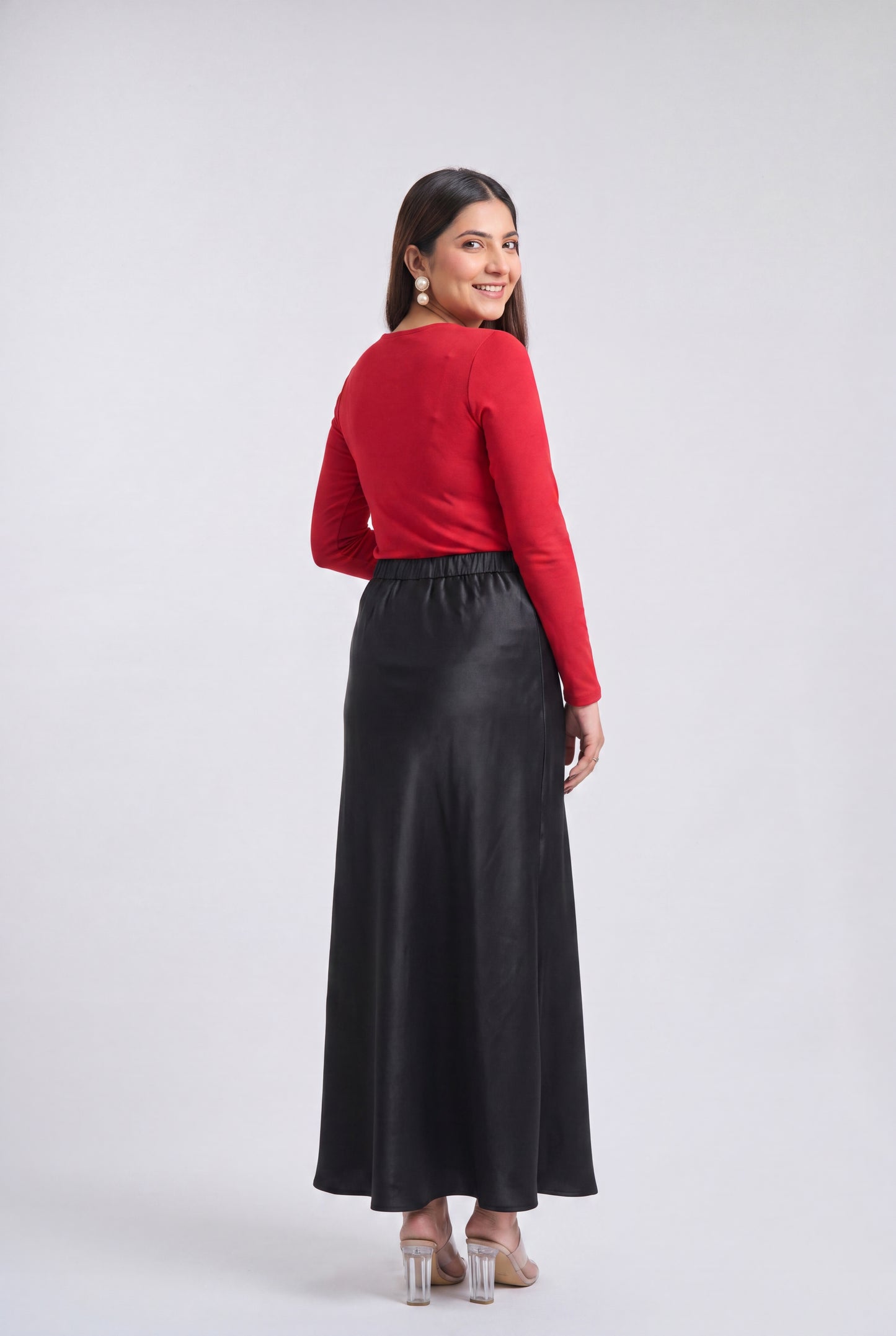 A woman is shown from the back wearing a bright red long-sleeve top and a long black skirt, standing against a light gray studio background.