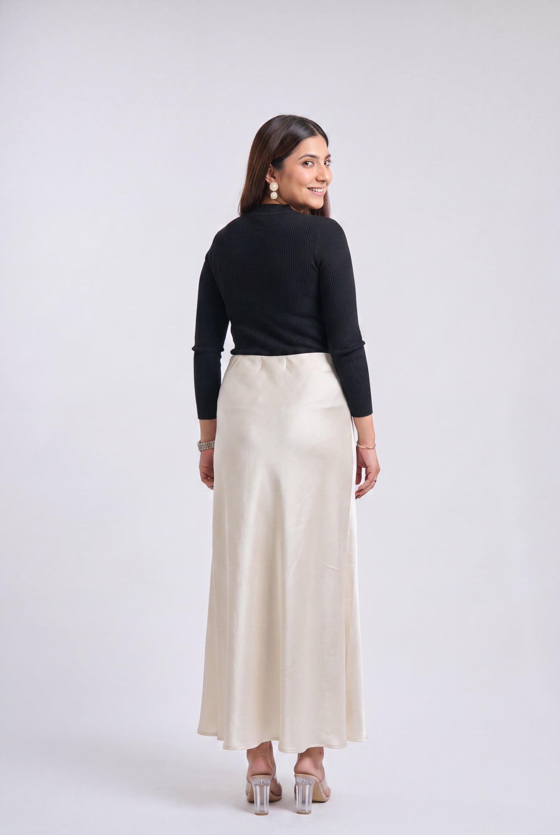 A woman is shown from the back wearing a black long-sleeve mock neck top and a high-waisted, floor-length cream satin skirt against a light gray studio background.