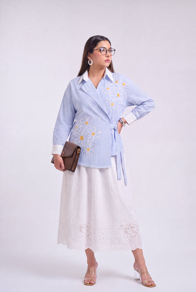 A woman models a light blue long-sleeve embroidered shirt with a white collar and tie waist, paired with a white midi skirt, holding a brown leather clutch against a light gray studio background.