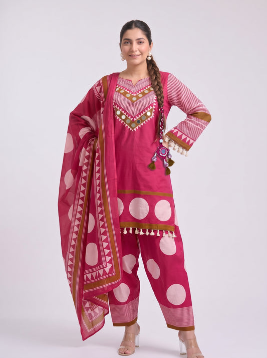 A woman modeling a pink traditional Indian salwar kameez suit with a matching dupatta. The outfit features white polka dots, gold and green geometric embroidery on the chest and sleeve cuffs, and gold fringe details.