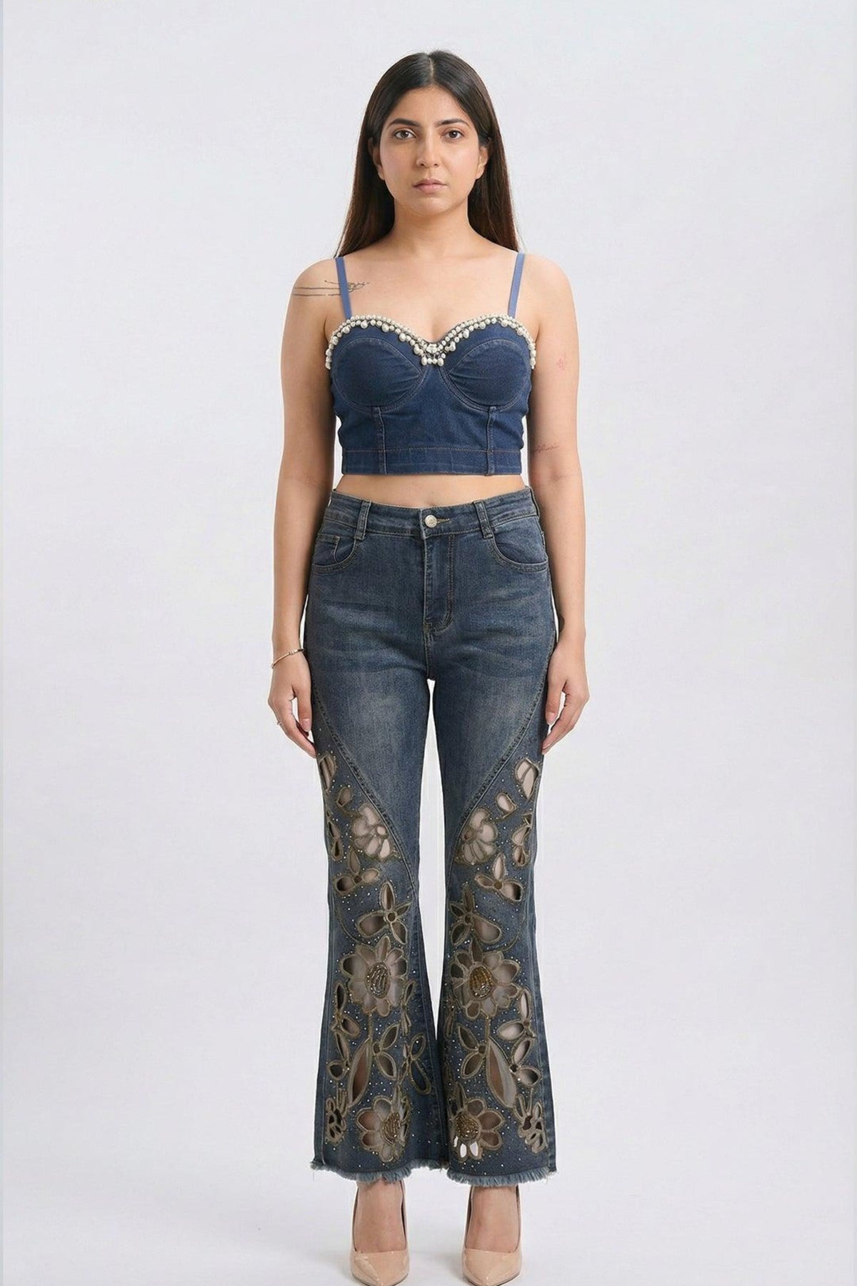 A model stands against a light gray studio background wearing a dark blue denim two-piece set consisting of a cropped camisole top with rhinestone trim and matching high-waisted flare jeans with gold floral embroidery on the lower legs.