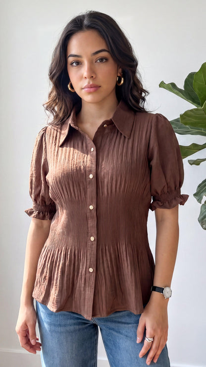 Woman wearing a brown button-up shirt and blue jeans against a white wall with a plant.