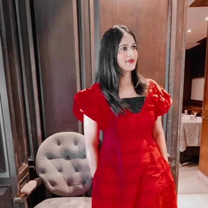 Woman in a red dress standing in an indoor setting with a chair and table in the background.