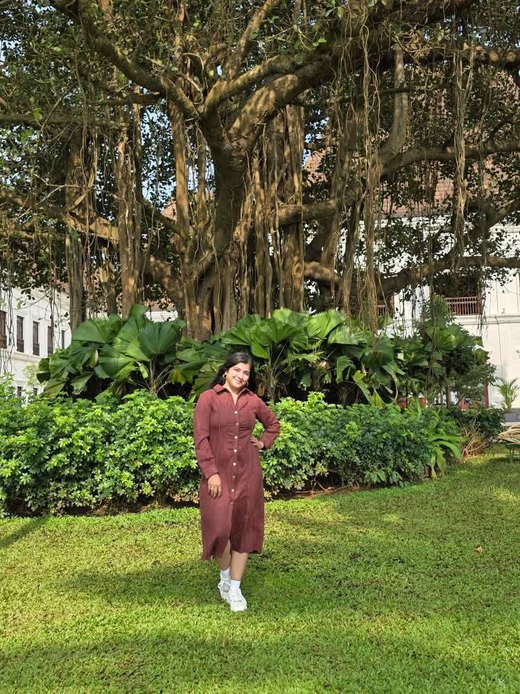 Woman in a red dress standing in a garden with large trees and greenery.