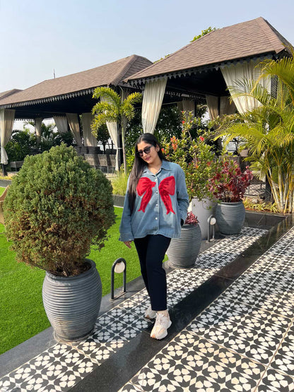Woman in a denim jacket with a red bow standing on a patterned patio with plants and a building in the background.