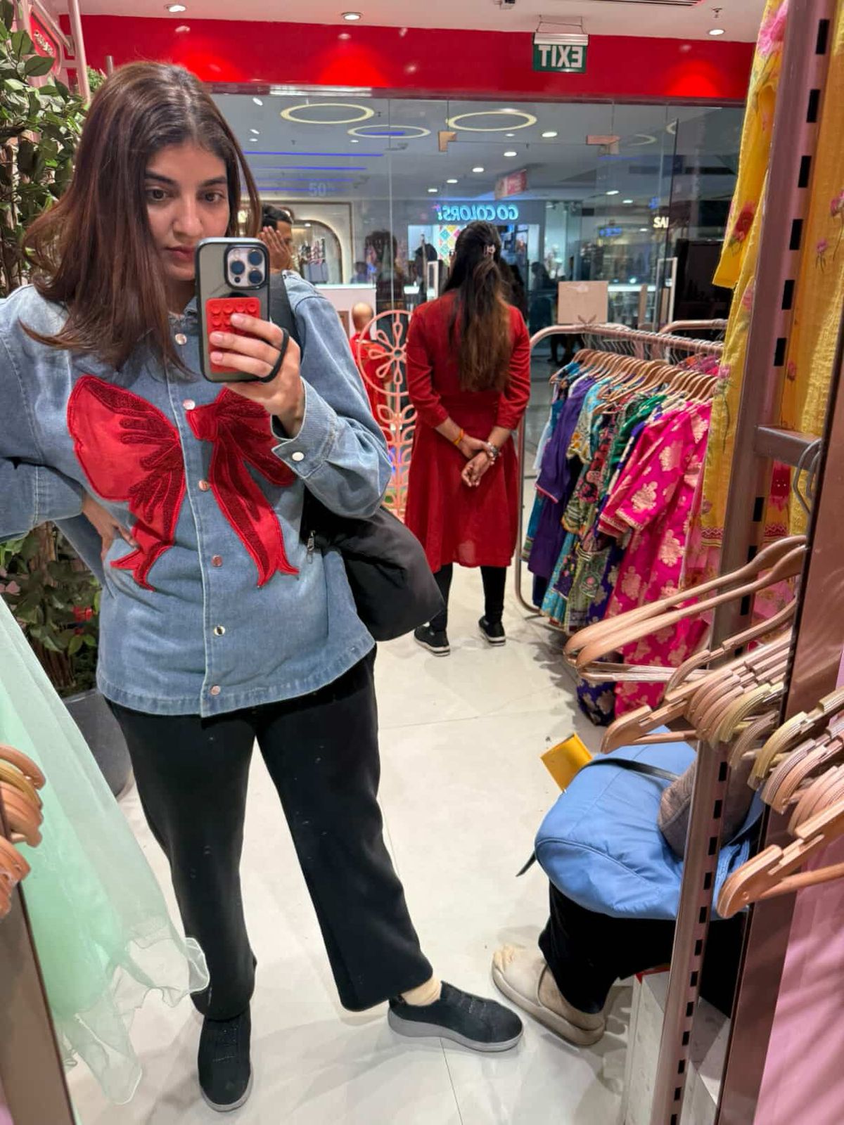 Woman taking a mirror selfie in a clothing store with racks of clothes and another person in the background.