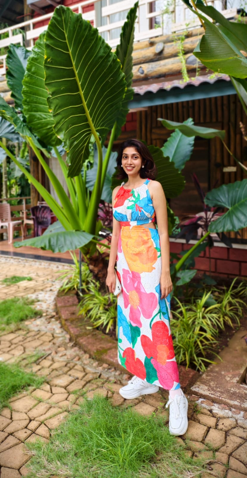 Woman in a colorful dress standing in a garden with large green leaves and a rustic building in the background.