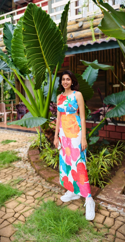 Woman in a colorful dress standing in a garden with large green leaves and a rustic building in the background.