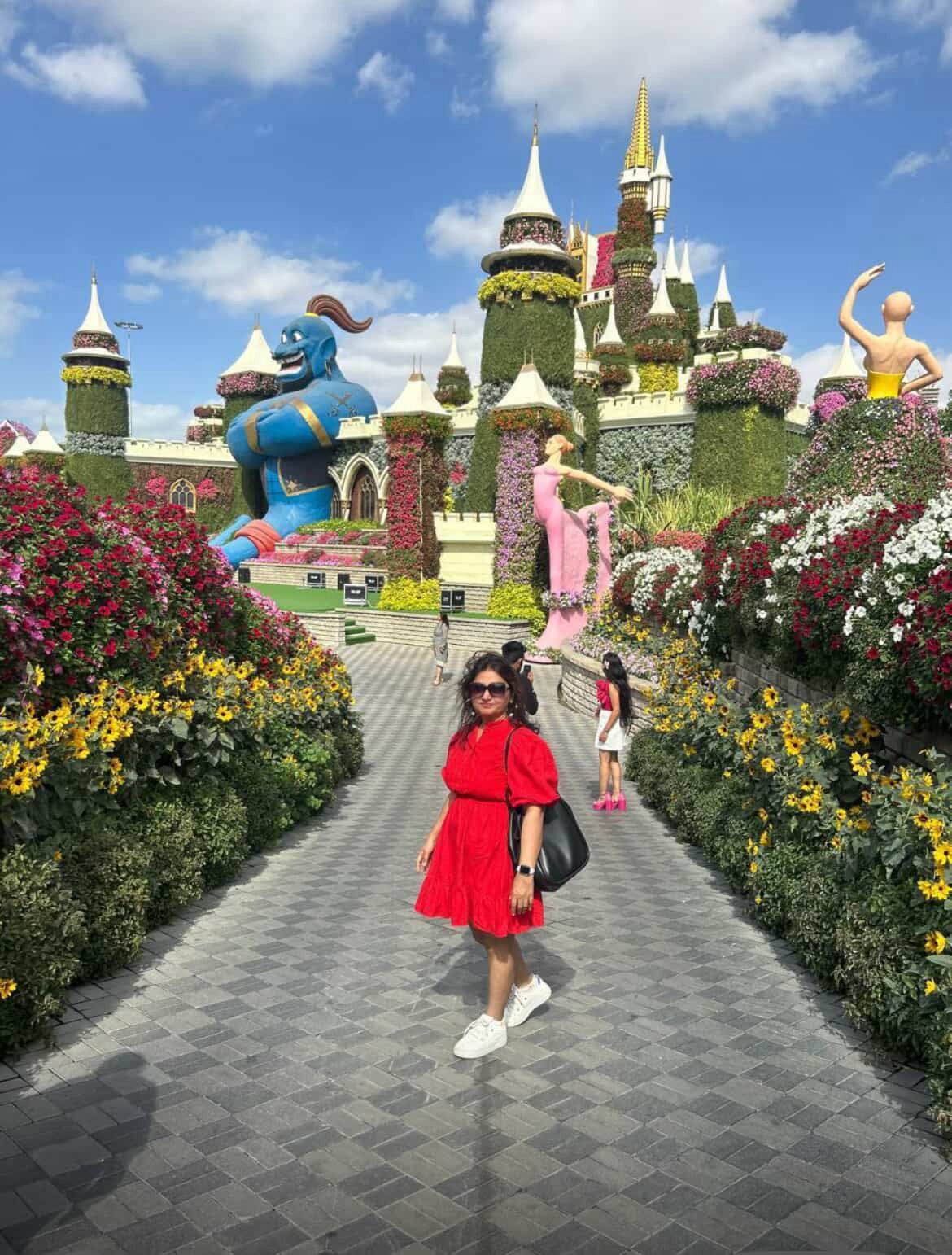 Woman in a red dress walking through a colorful garden with a castle-like structure.