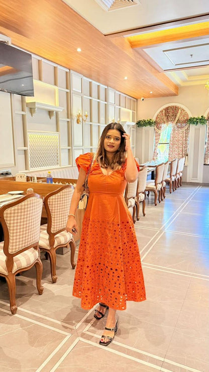 Woman in an orange dress standing in a restaurant with wooden chairs and tables.