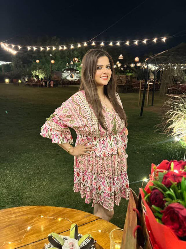 Woman in a patterned dress standing outdoors at night with string lights and a table with flowers.