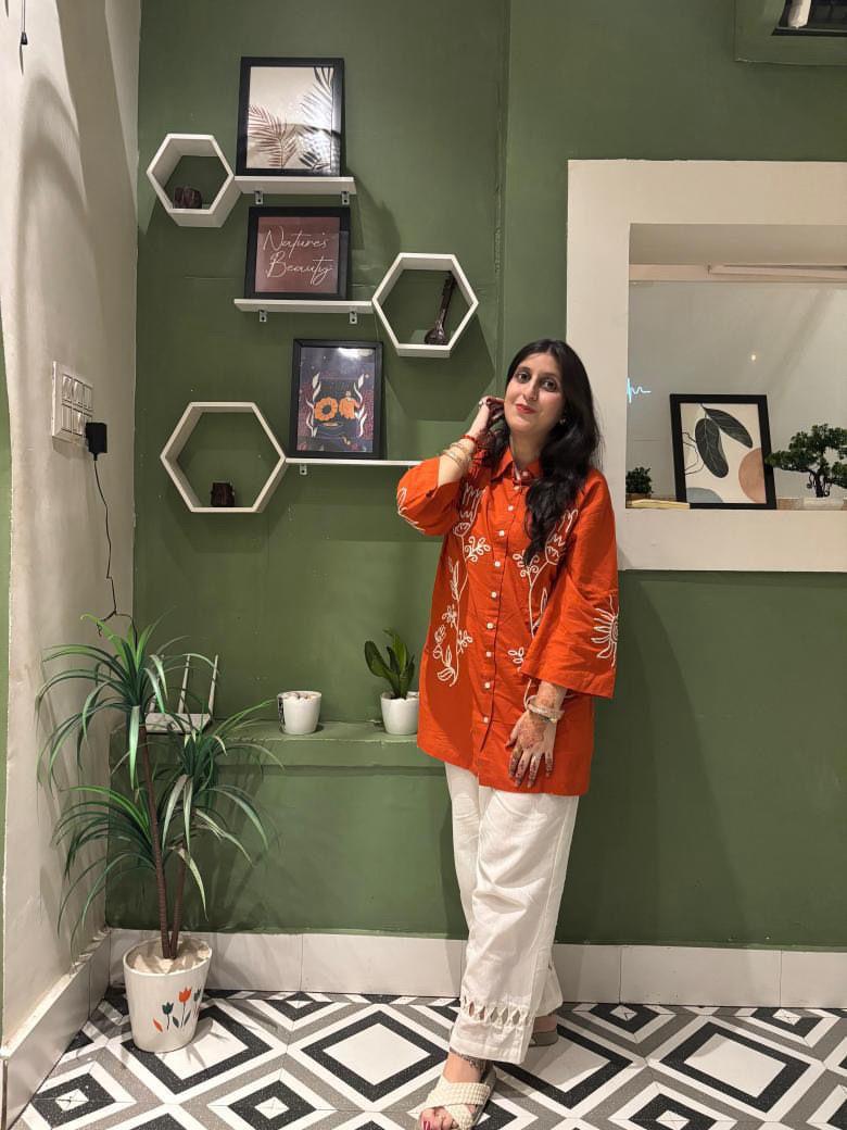 Woman in an orange shirt and white pants standing in a room with green walls and geometric wall shelves.
