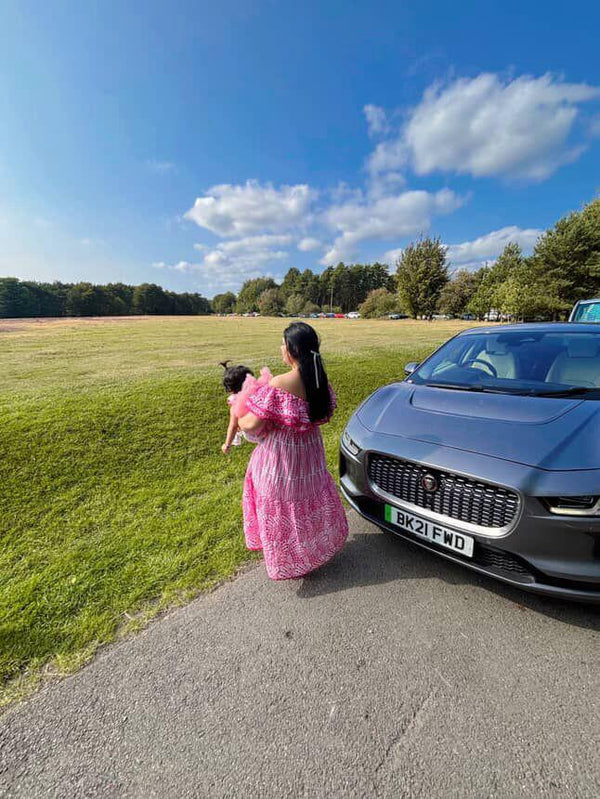 Woman in a pink dress holding a child next to a blue car on a sunny day.