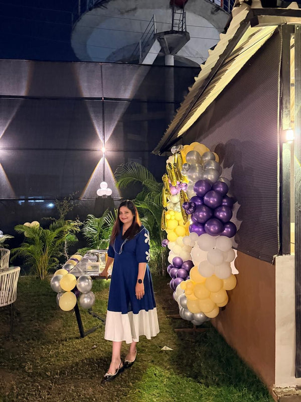Woman in a blue dress standing next to a balloon arch with yellow, purple, and white balloons at night.