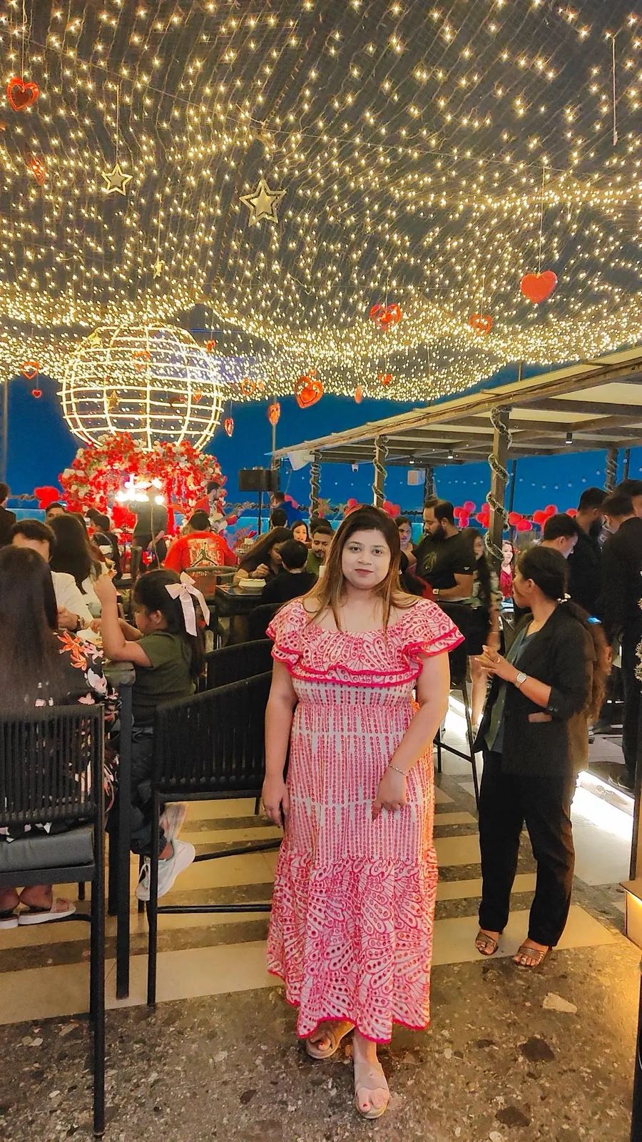 Woman in a pink dress standing in a festive outdoor setting with lights and decorations.