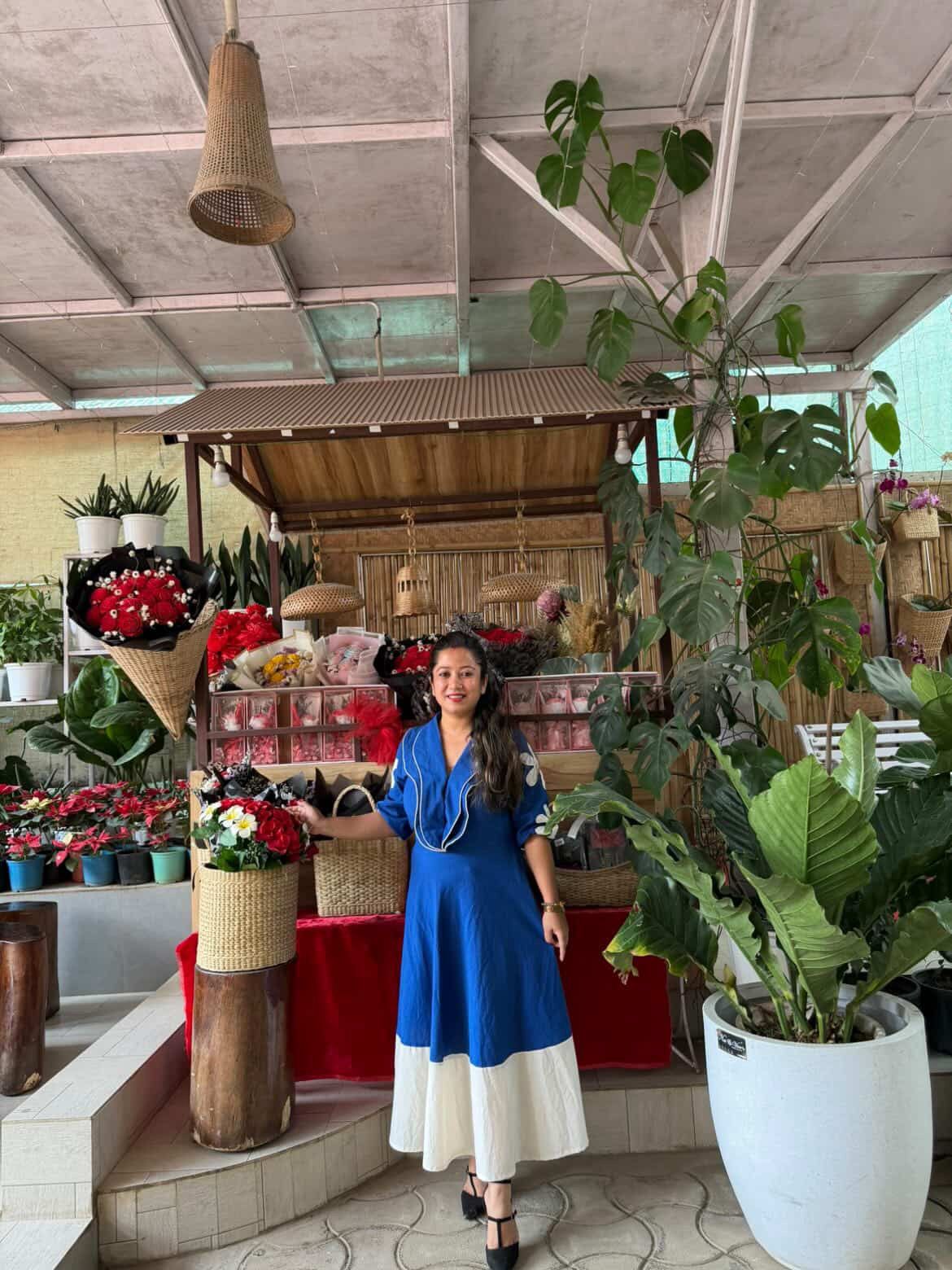 Woman in a blue dress standing in a floral shop with plants and flowers around.