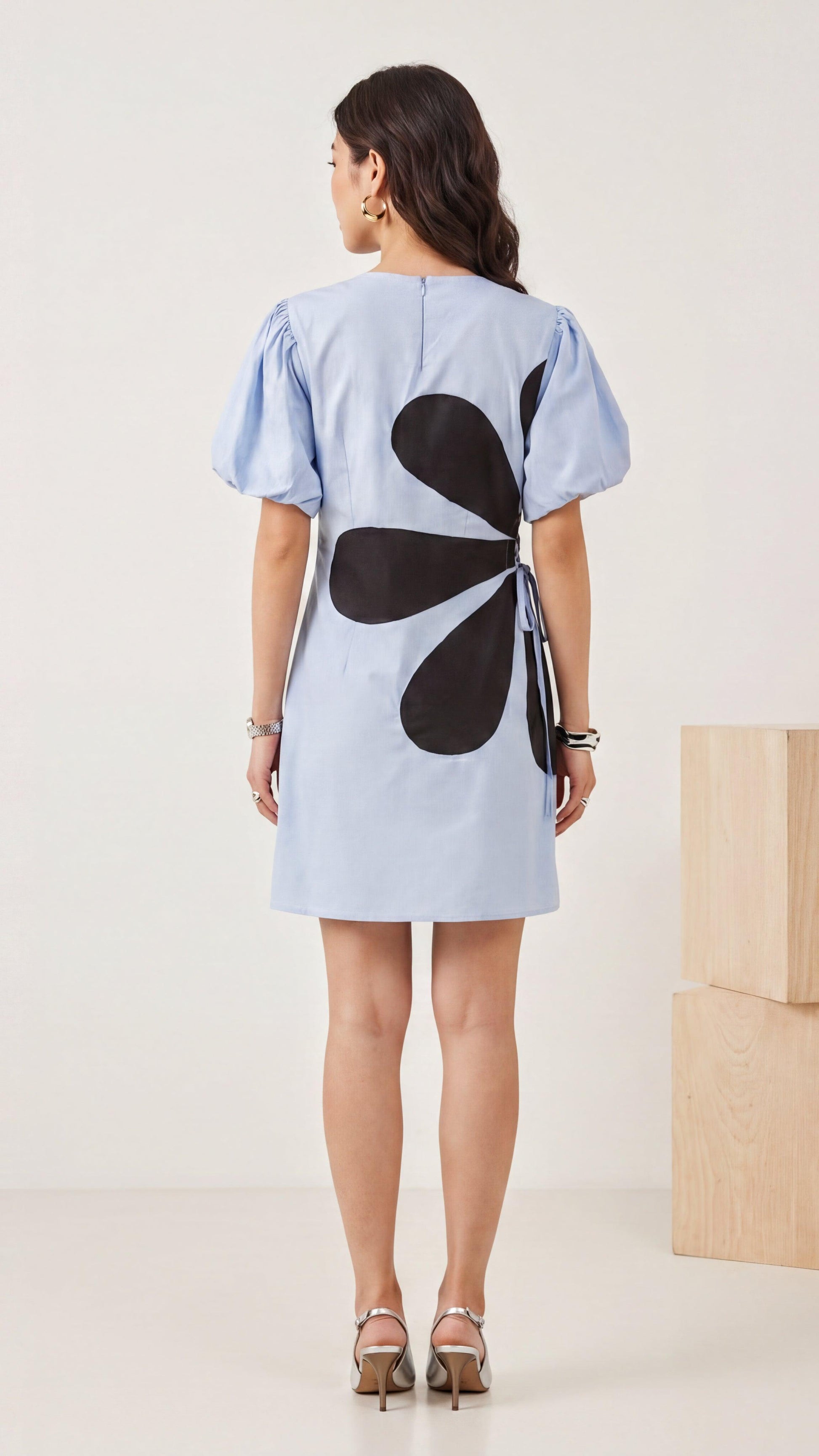 Woman wearing a light blue dress with a black floral design on the back, standing against a white background.