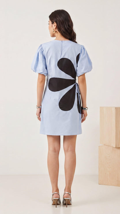 Woman wearing a light blue dress with a black floral design on the back, standing against a white background.
