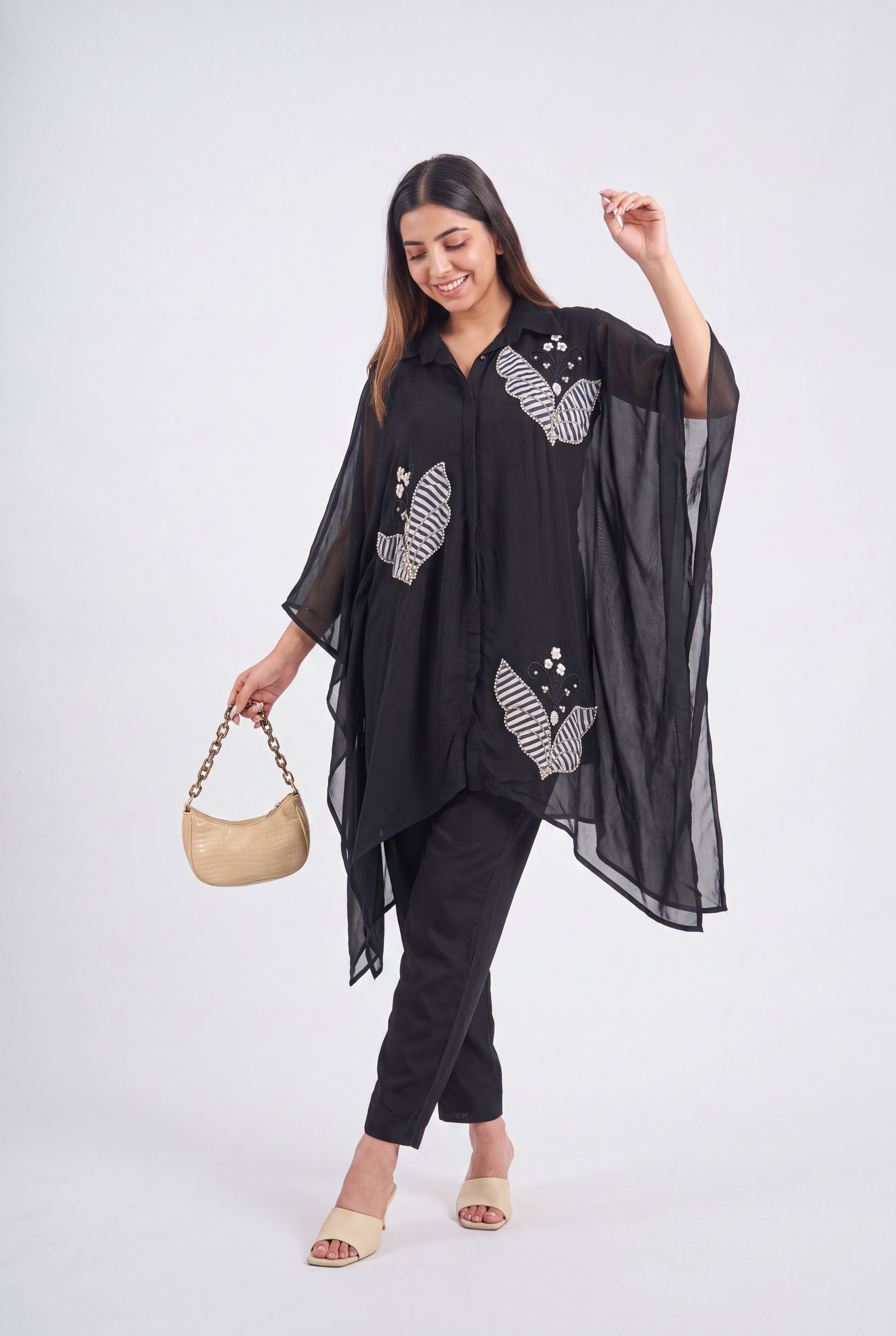 A woman modeling a black sheer long-sleeve tunic top with white and gray zebra-patterned embroidery, paired with black pants and beige sandals, holding a beige handbag against a light gray studio background.